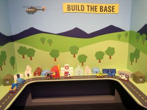 Photo of a miniature army base with small buildings lined alongside a road with plastic cars. In the background is a cartoon scene of trees, grass, and mountains with a helicopter flying above trailing a banner that says “build a base.”