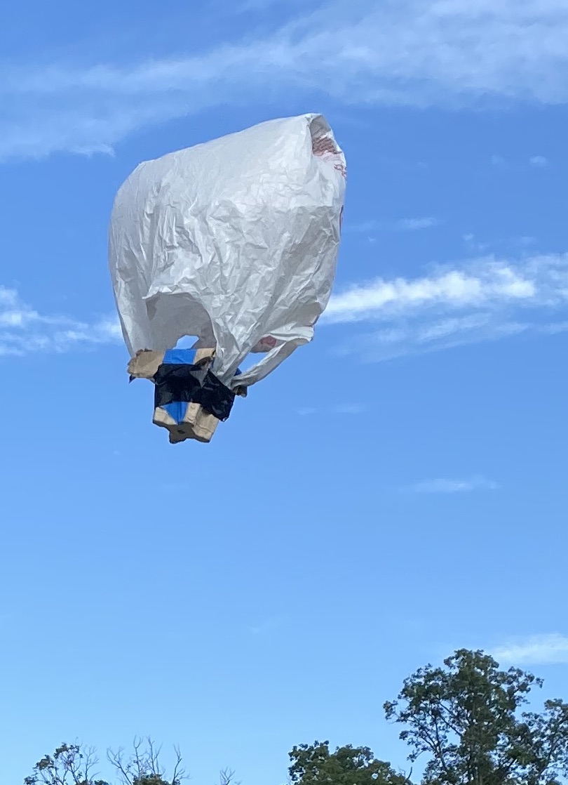 Photo of science project landing capsule. Close up a newspaper-wrapped box with an attached plastic bag parachute. The newspaper is folded around a box. The bag is attached to the box wit rubber bands wrapped around the box.