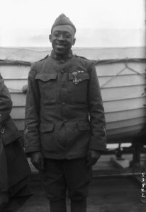 Black and white photo of an African American Soldier in uniform on the deck of a ship in front of a life boat. 