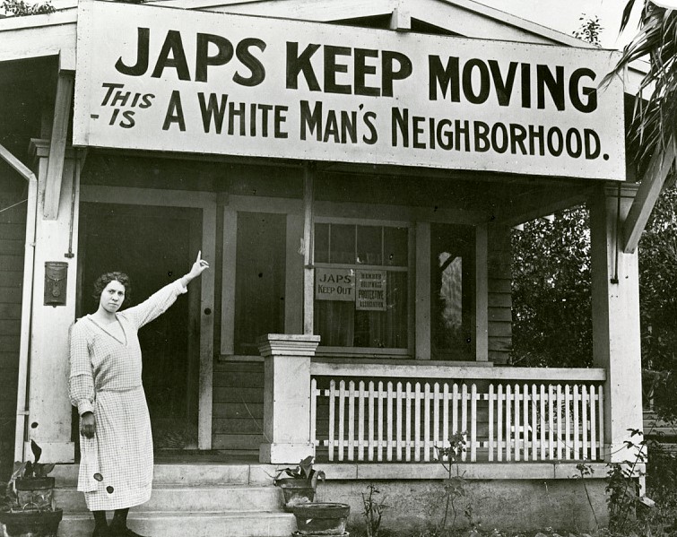 White woman points at a large sign on the house behind her that says "Japs keep moving - this is a white man's neighborhood."