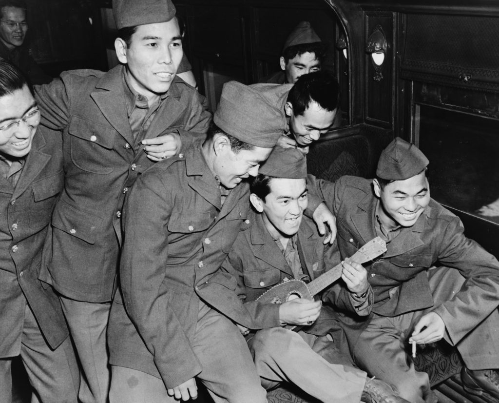 Group of Japanese American Soldiers gather around another soldier with a ukulele. 