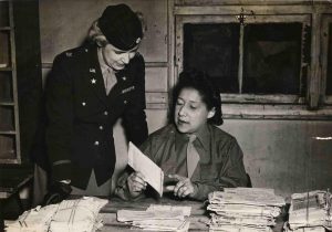 Alyce Dixon in uniform sits at a desk piled with mail. A white female officer stands over her as they discuss a piece of mail.