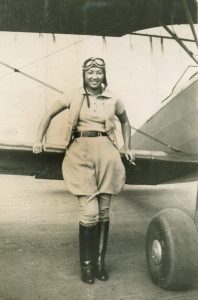 Hazel Ying Lee in aviator outfit and helmet standing next to an airplane