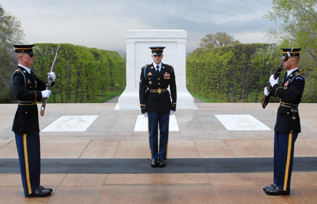 Three Soldiers in dress blue uniforms stand at attention in front of the Tomb. Two Soldiers face each other holding guns across their chests.
