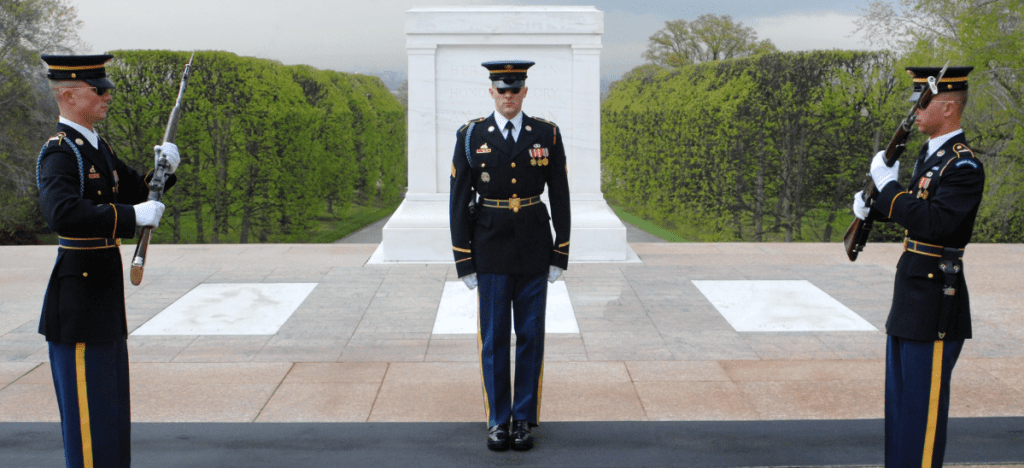 Three Soldiers in dress blue uniforms stand at attention in front of the Tomb. Two Soldiers face each other holding guns across their chests.