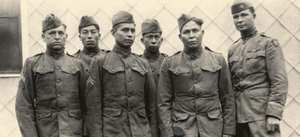 6 Native American men in World War I era Army uniforms stand in front of a building. 