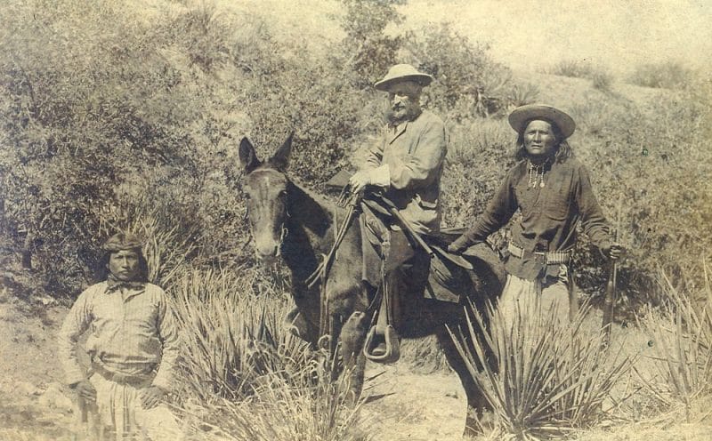 On left, a Native American man stands near a horse with an Soldier mounted on the horse. Another Native American man stands to the right of the horse. The men are surrounded by brush. 