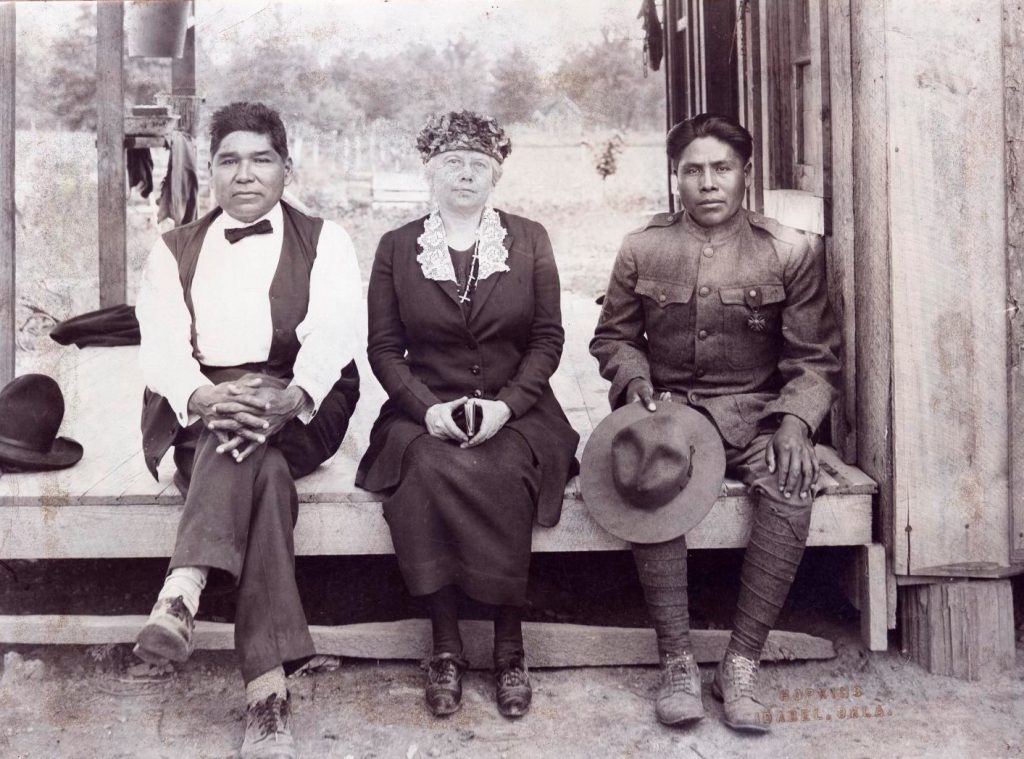 From left, Native American man in vest and bowtie with woman in a dress sit next to Native American Soldier in uniform