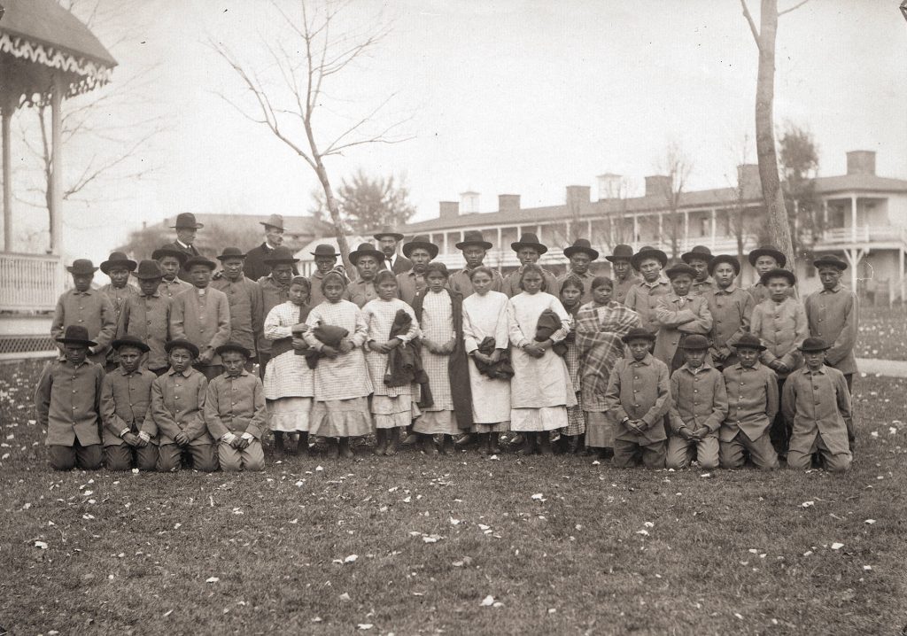 Group of Native American children in posed in Western clothing. The boys wear pants, a buttoned jacket, and a brimmed hat. Girls wear long dresses and hold blankets. Three white men stand behind them.