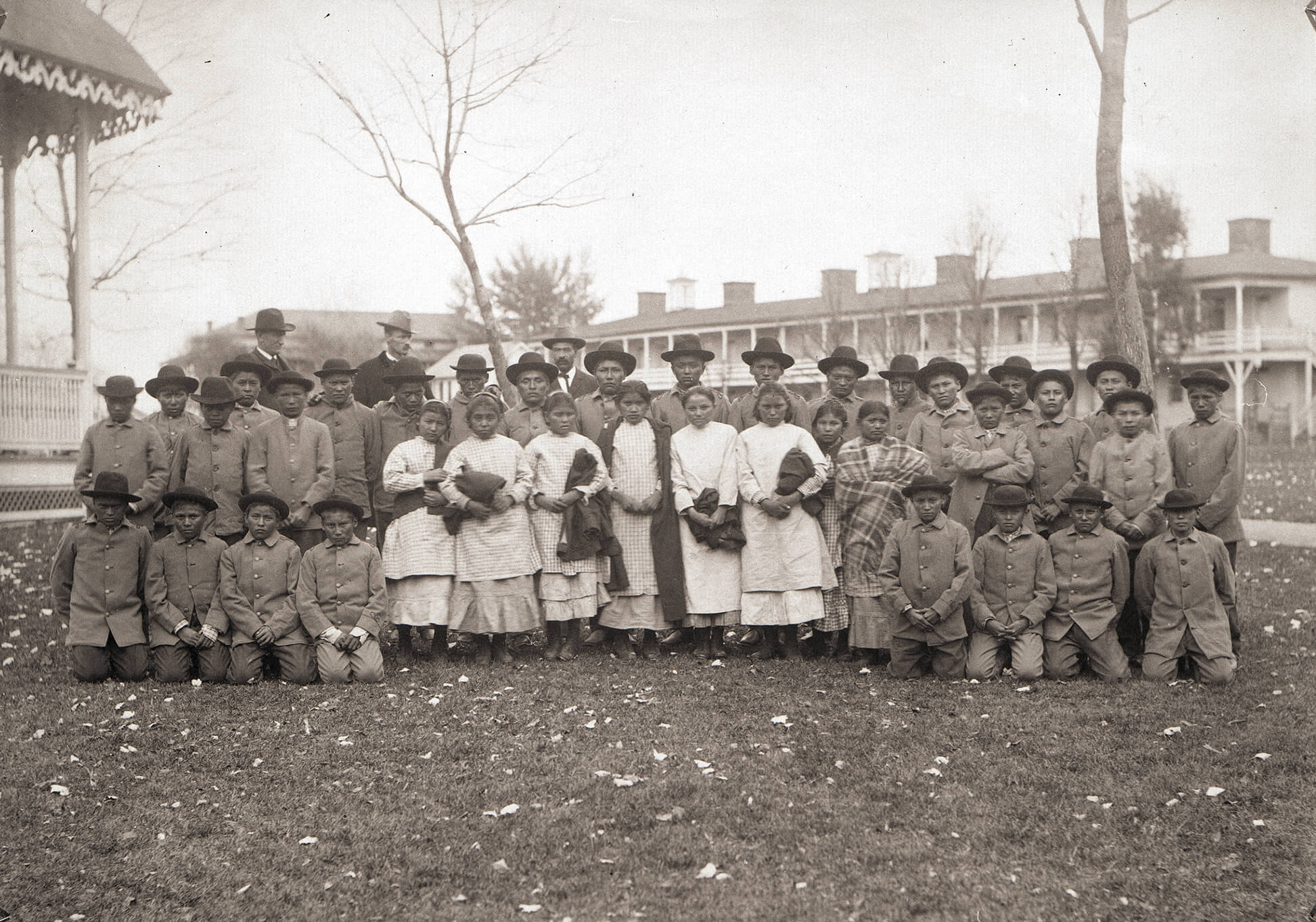 Group of Native American children in posed in Western clothing. Three white men stand behind them.