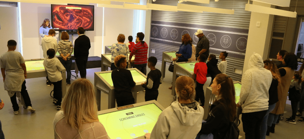 people standing at touch screen kiosks with a leader at the front of the room. The screen at the front of the room shows a map of an island that is glowing red.