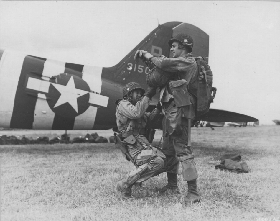 Two Soldiers in front of an airplane. One Soldier inspects the gear of the other soldier. 