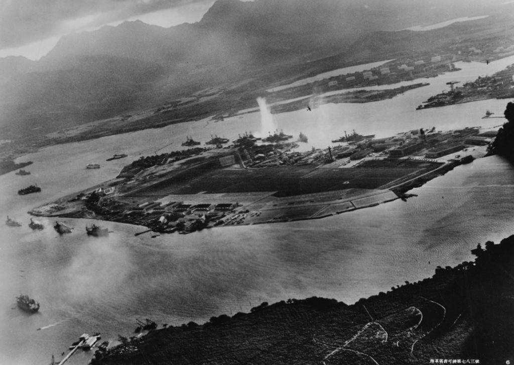 Aerial view of an island with battleships. A plume of water erupts into the air next to one of the ships.