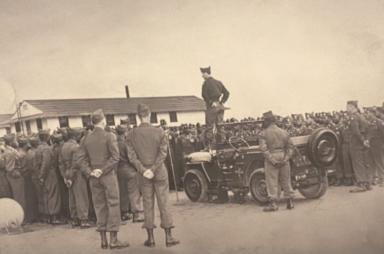 A Soldier stands on the hood of a jeep surrounded by a formation of mostly Black Soldiers