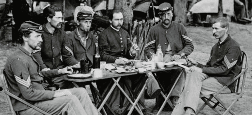 6 Civil war soldiers sit around a table filled with food and drinks