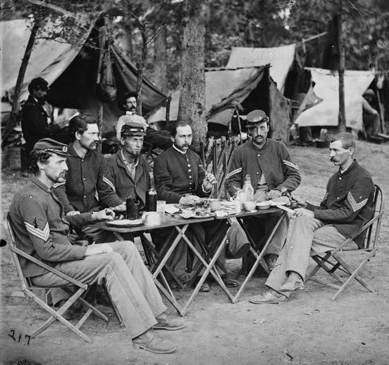 6 Soldiers sit around a table filled with food and drink. Tents and trees are in the background