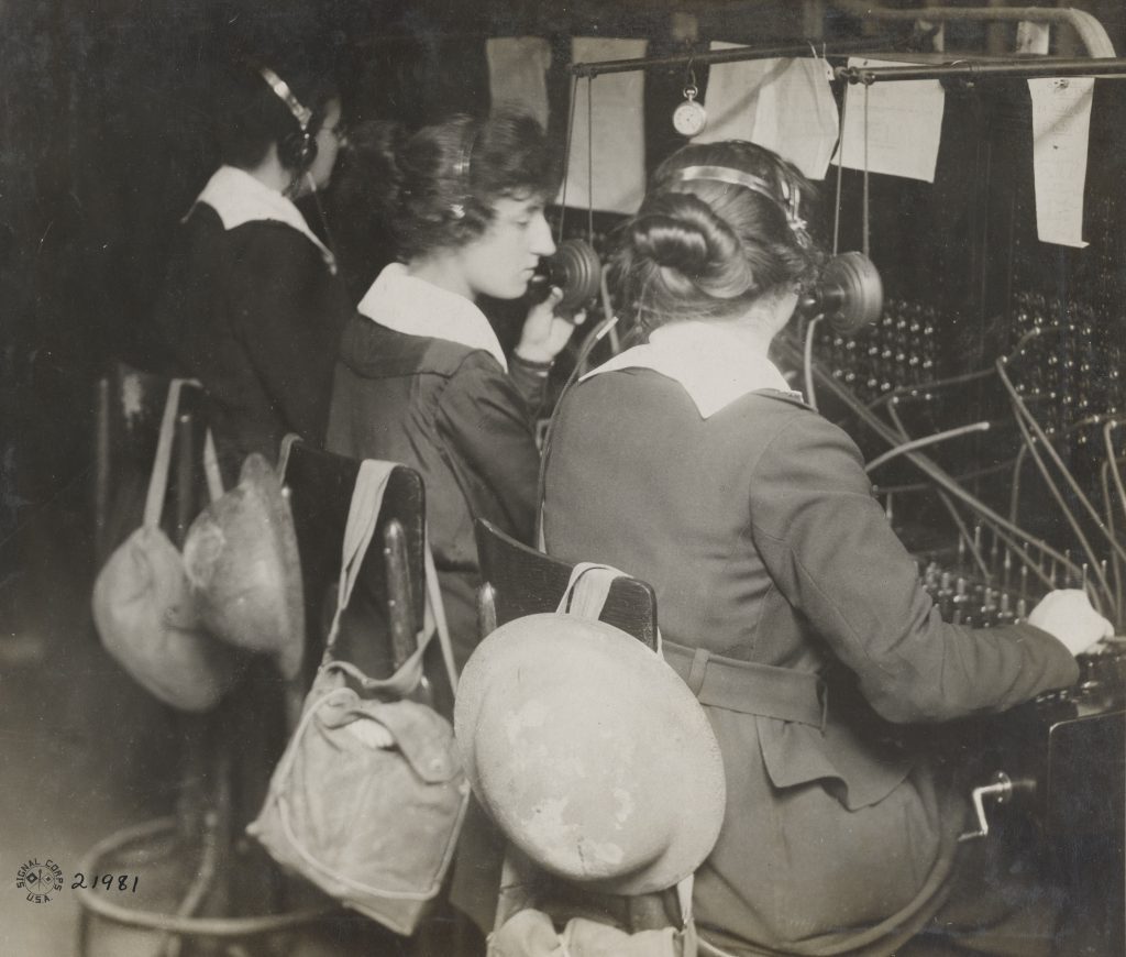 Three women with headphones sit at a switchboard with helmets and gas masks slung on the back of their chairs. 