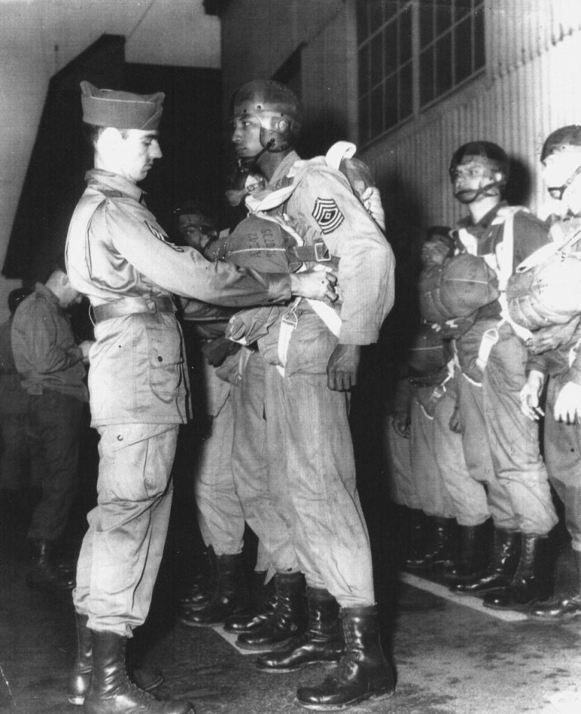 a white master sergeant inspects the kit on a Black first sergeant as he prepares for a jump. Other white Soldiers are similarly lined up nearby 