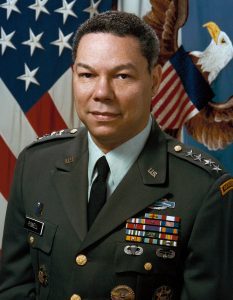 General Colin Powel in uniform in front of a US flag and the flag of the chairman of the joint chiefs of staff featuring an eagle with a shield.