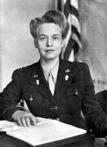 Oveta Culp Hobby seating at a desk with her right hand resting on an open book with an american flag in the background.