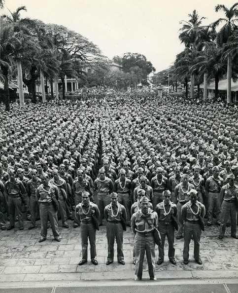 Large group of uniformed Japanese American men standing outside in formation with palm trees on either side. 