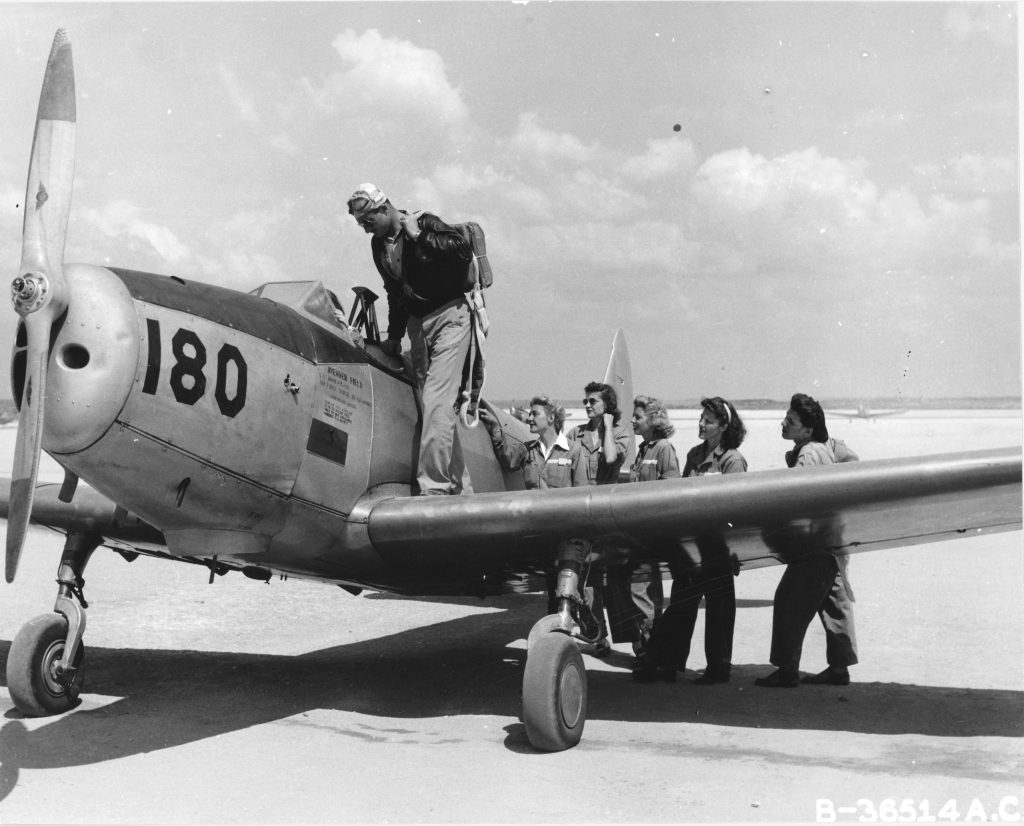 5 women in flight uniforms stand on the ground next to the wing of a single engine propeller plane. A male pilot stands on the wing while another woman sits in the cockpit.