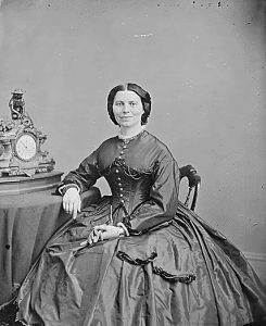 Clara Barton seated at a table holding a pair of gloves in her left hand. Her right arm rests on the table near a clock. 