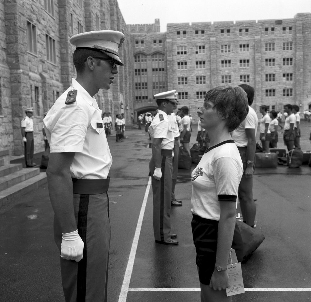 In the foreground, a male cadet in uniform stands facing a female cadet in an exercise uniform. In the background, other new cadets and cadets in uniform stand in the courtyard.