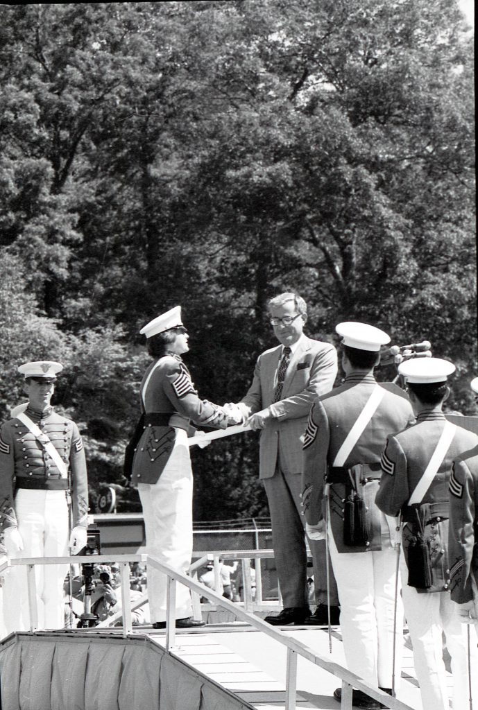 Andrea Hollen, in her West Point uniform, stands on stage, receives her diploma and shakes a man's hand while other cadets line up on either side of the stage.