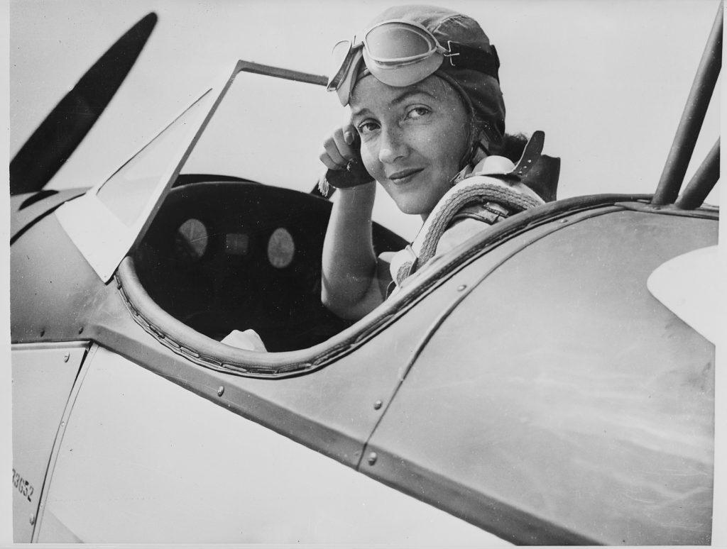 woman sits in the open cockpit of a propeller airplane. She is wearing a flightcap and goggles and holding on to one side of the flight cap