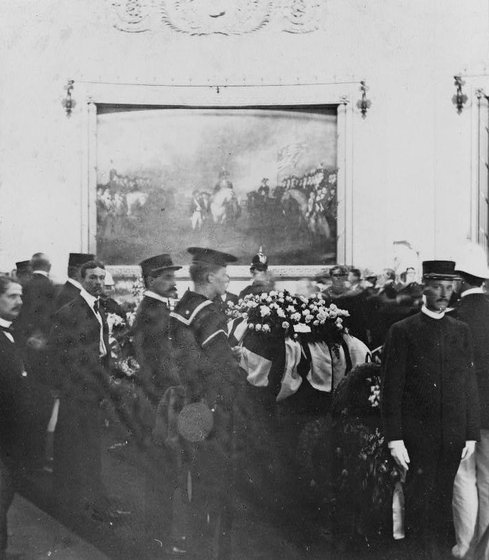 Casket covered with flag and flowers lays in the middle of a crowd of men with painting of George Washington on his horse in the background.