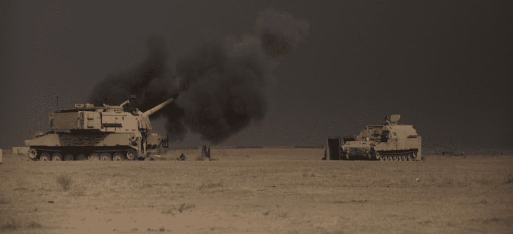 two tanks sit in a desert at dusk. the left tank is surrounded by a cloud of smoke from their weapon. 
