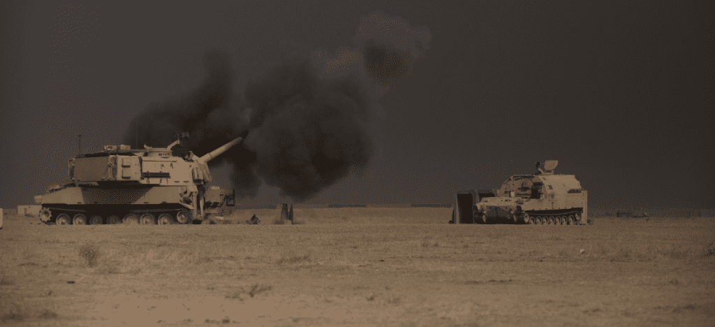 two tanks sit in a desert at dusk. the left tank is surrounded by a cloud of smoke from their weapon.