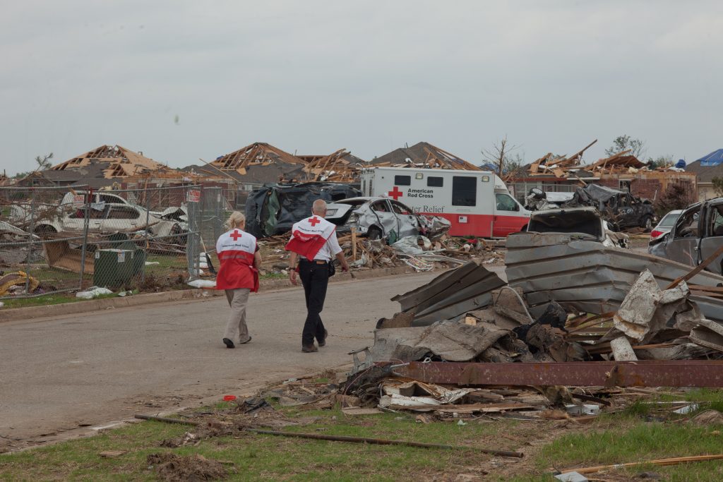 A man and woman wearing Red Cross jackets walk down a street strewn with totaled cars and building material. Shattered houses and a Red Cross disaster Relief vehicle are in the background.