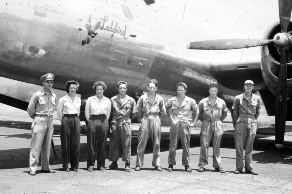 6 men and 2 women, all in uniform, stand in front of a large plane. The name "Ladybird" is painted on the side of the plane alongside a cartoon "female gremlin" dressed as a pilot.