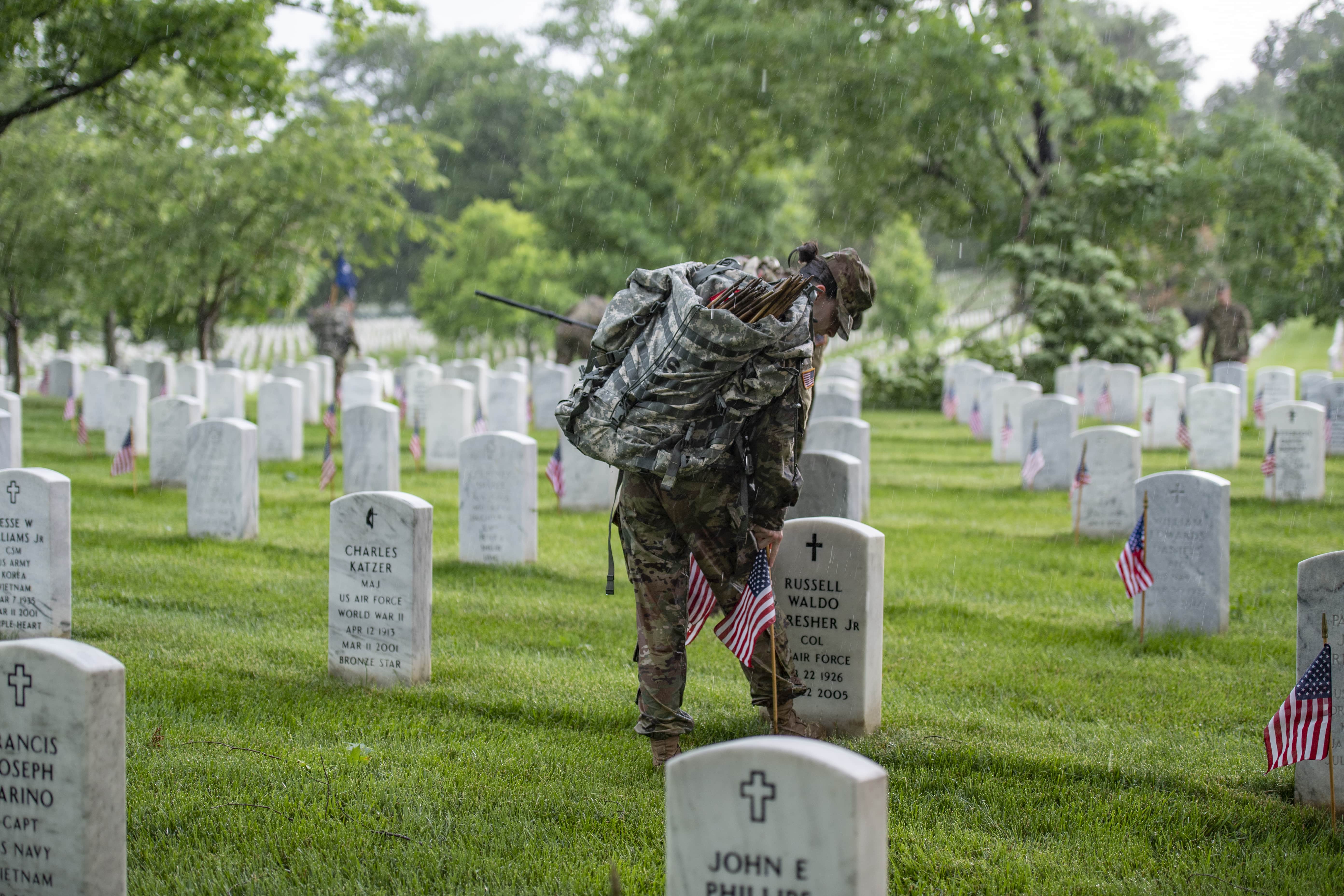 A female Soldier carrying a backpack of flags places an american flag a bootlength away from a gravestone. 