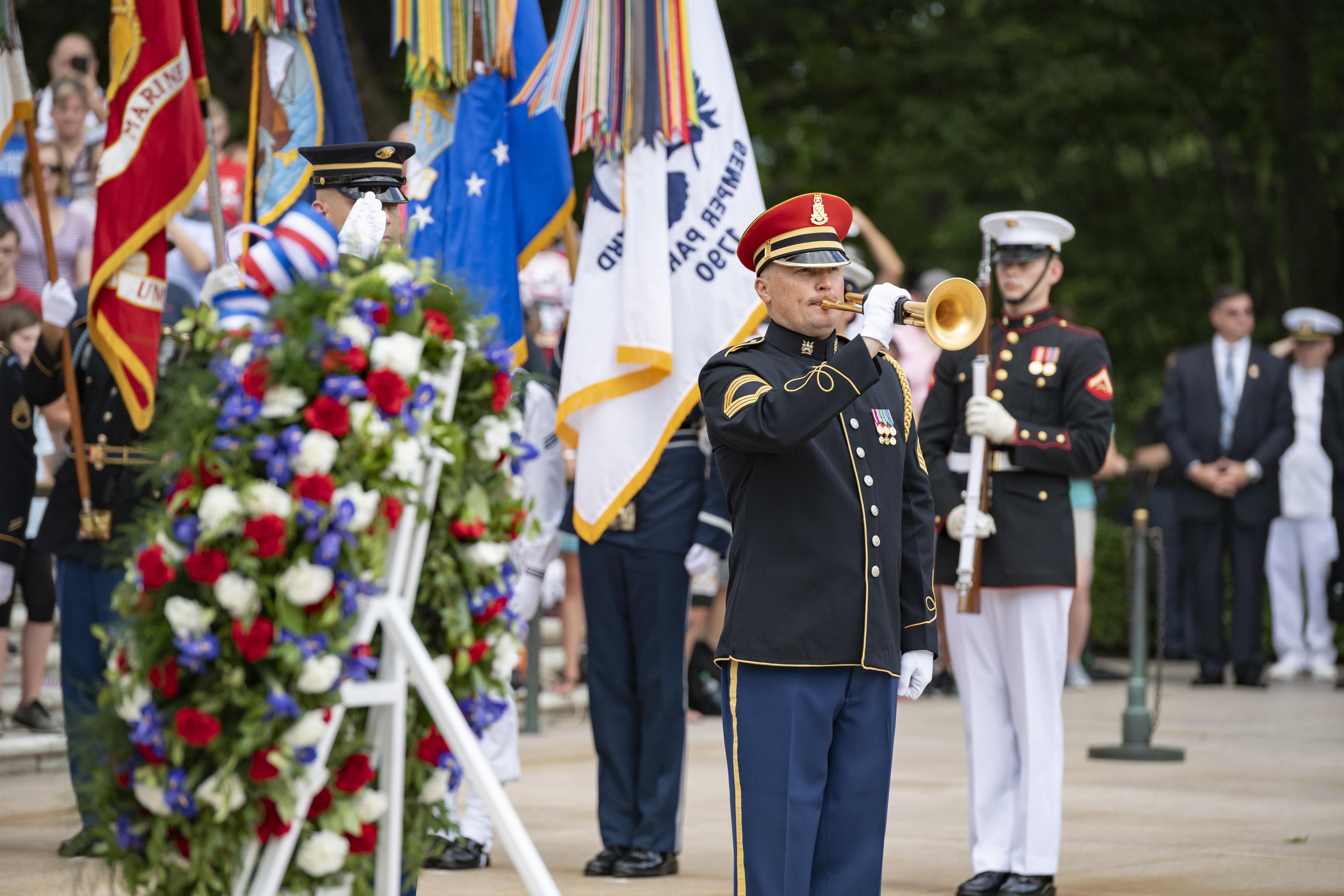 A Soldier in dress uniform plays the bugle. A flower wreath in the foreground and the flags of other service branches in the background. 