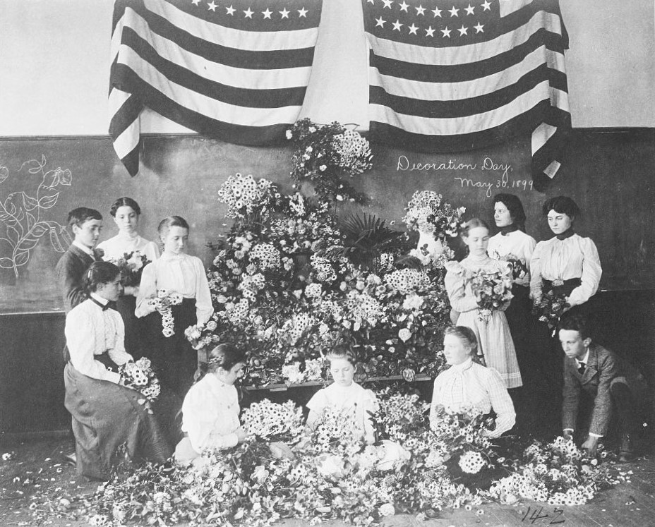girls and boys gather in front of a chalk board surrounded by flowers. Two American flags hang above the chalk board.