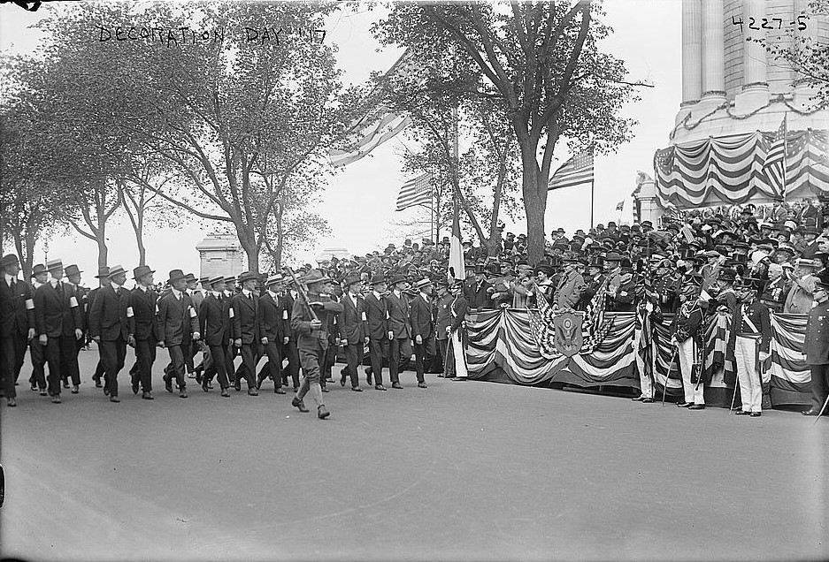 A uniformed Soldier carrying a rifle on his shoulder leads a column of men in suits and armbands down a street. Crowds gather on the side. A patriotically decorated viewing booth is in the background.