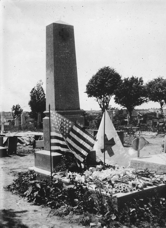 obelisk grave stone surrounded by flowers with an American flag and Red Cross flag. The rest of the cemetery is in the background