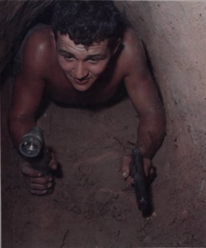 Shirtless young man crawls on his stomach through a small tunnel holding a flashlight in his right hand and a pistol in his left hand. 