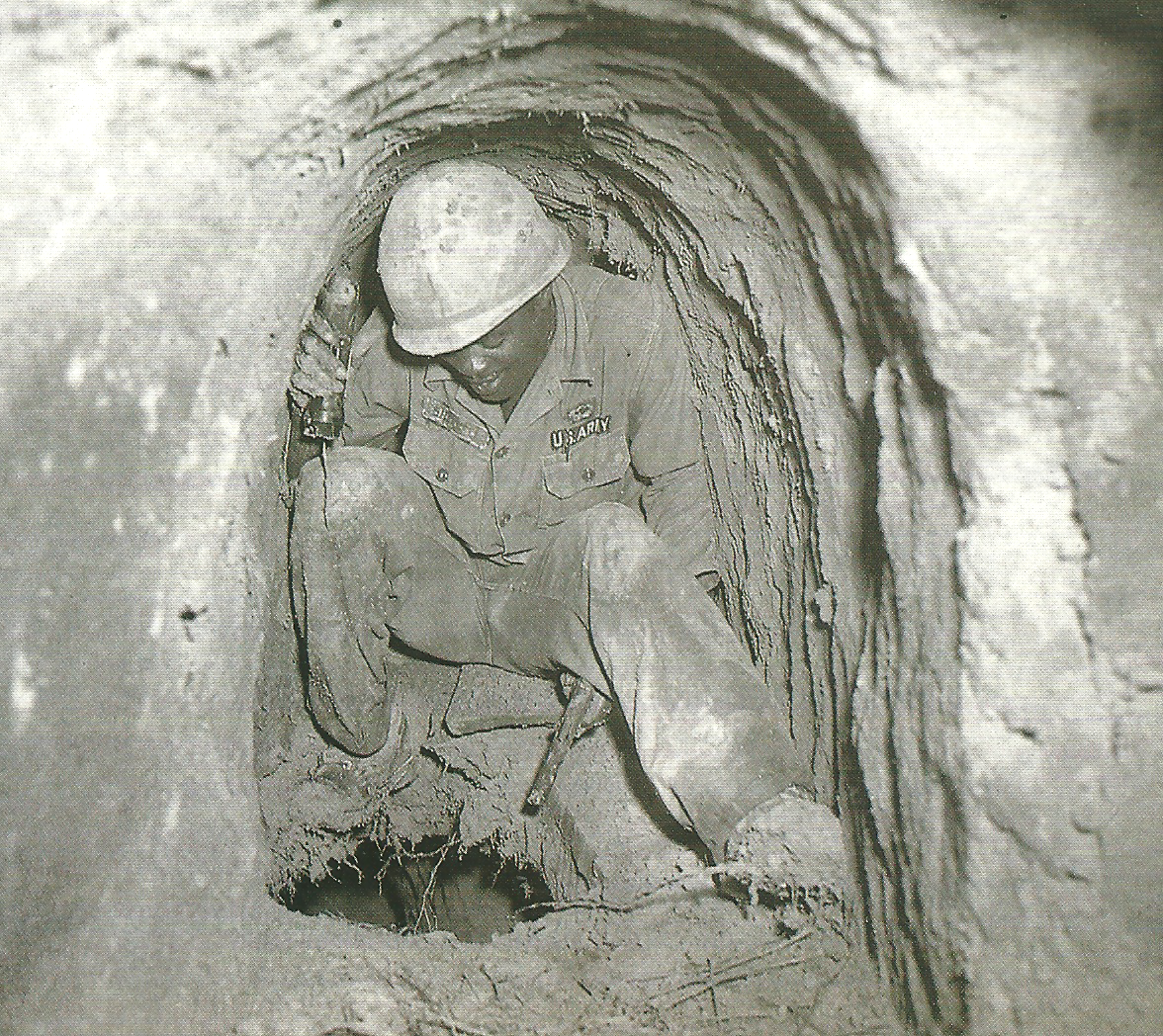 Man in uniform huddled in a tunnel over a hole in the ground holding a flashlight in his left hand and a pistol in his right hand, both pointed at the hole.