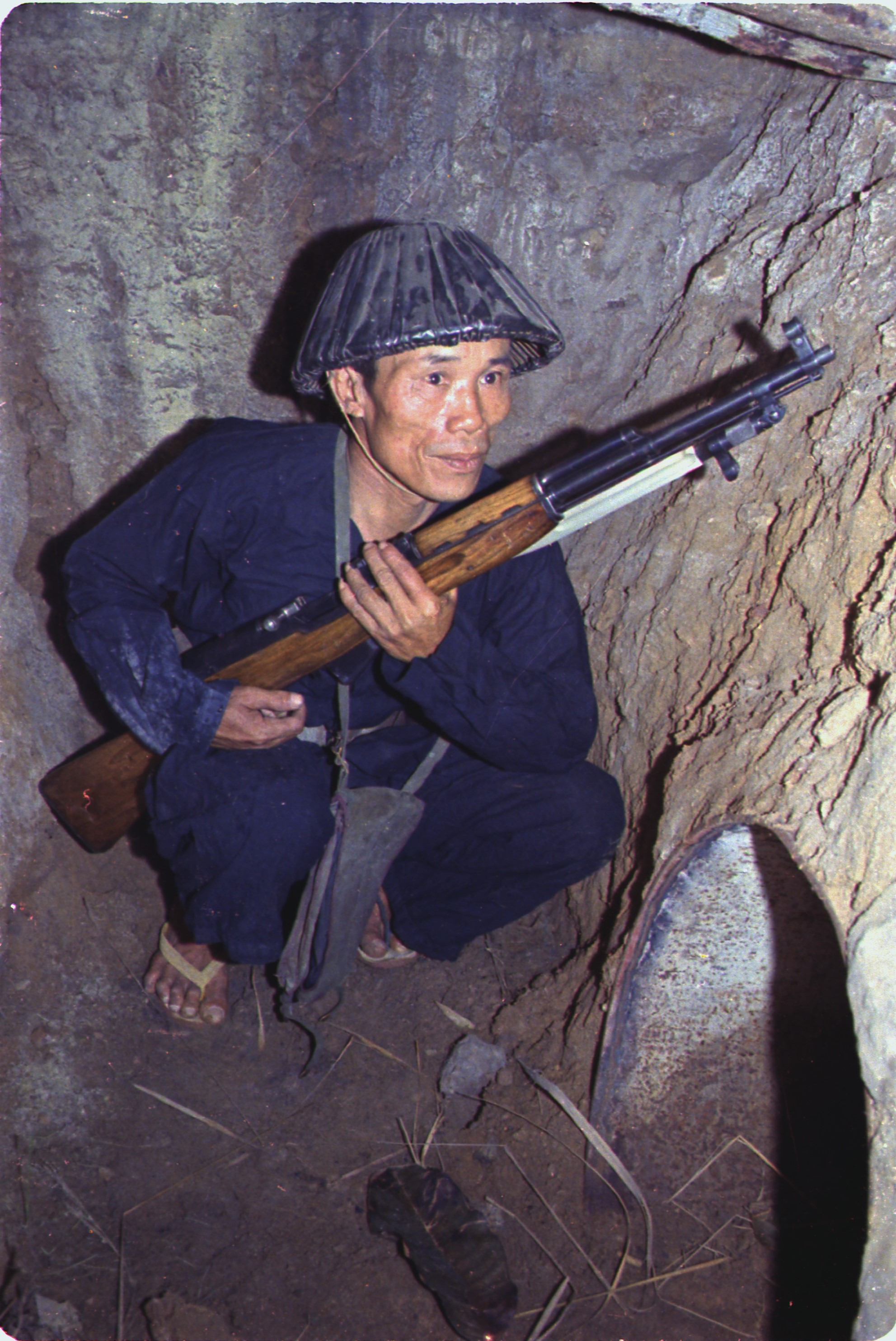 Viet Cong soldier crouched in a tunnel holding a rifle outside the entrance to a smaller tunnel. 
