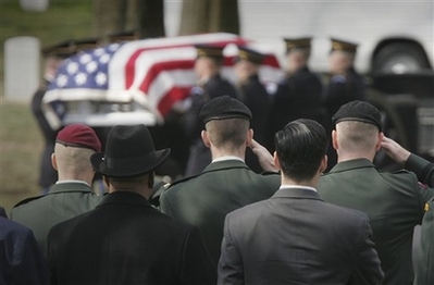 Men in uniform and civilian dress face away from the camera looking at an American Flag covered casket in the background.