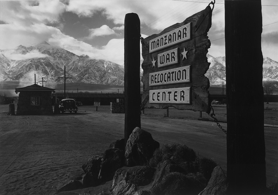 Wooden sign with the words "Manzanar *War* Relocation Center". To the left, a car sits at a guard house. Mountains in the background. 