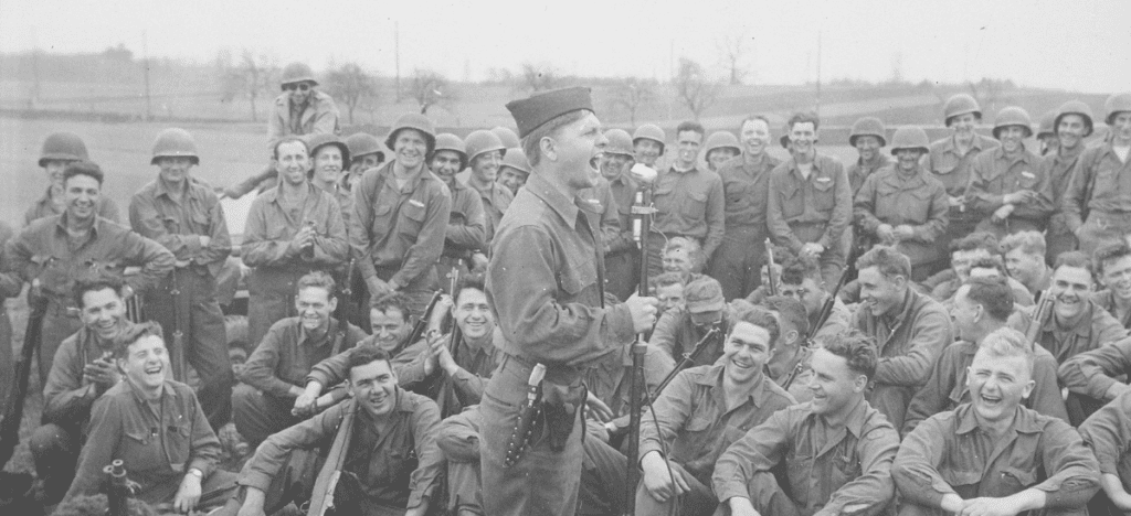 Mickey Rooney, in uniform with a knife on his waistband, stands at a microphone with his mouth open. Soldiers surround him, laughing. 