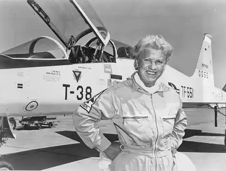 Jackie Cochran in flight suit wit her hands on her hips standing in front of a jet plane with the cockpit open. 