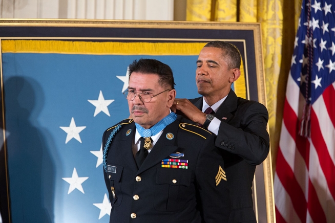 President Obama stands behind Sgt. Erevia. Obama hooks the medal of honor around Erevia's neck. The Medal of honor flag and the American flag are behind the men.