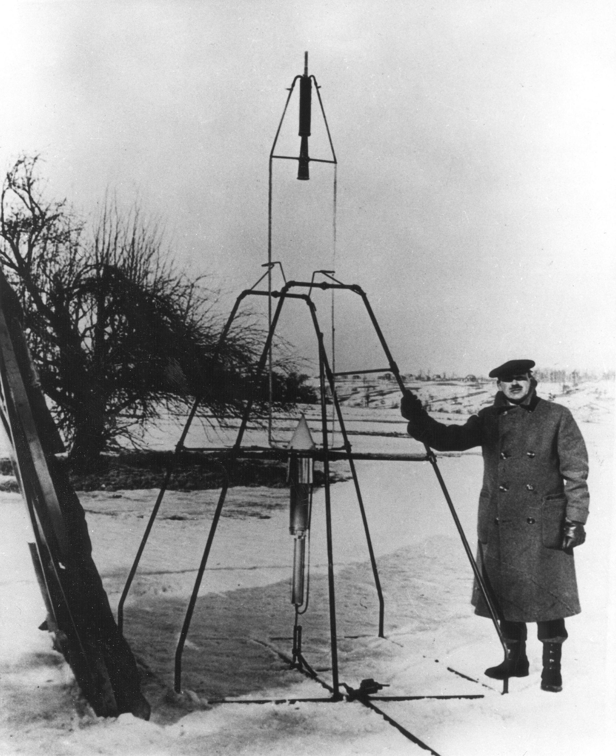 Goddard stands in the snow next to a structure holding his rocket. The structure and rocket are about twice as tall as he is. 