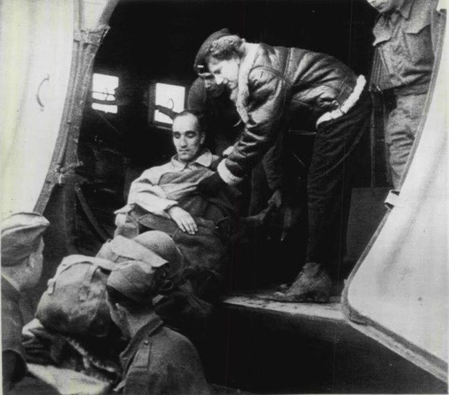 Aleda Lutz in flight jacket hands a wounded soldier on a stretcher out of the plane to two soldiers on the ground. 